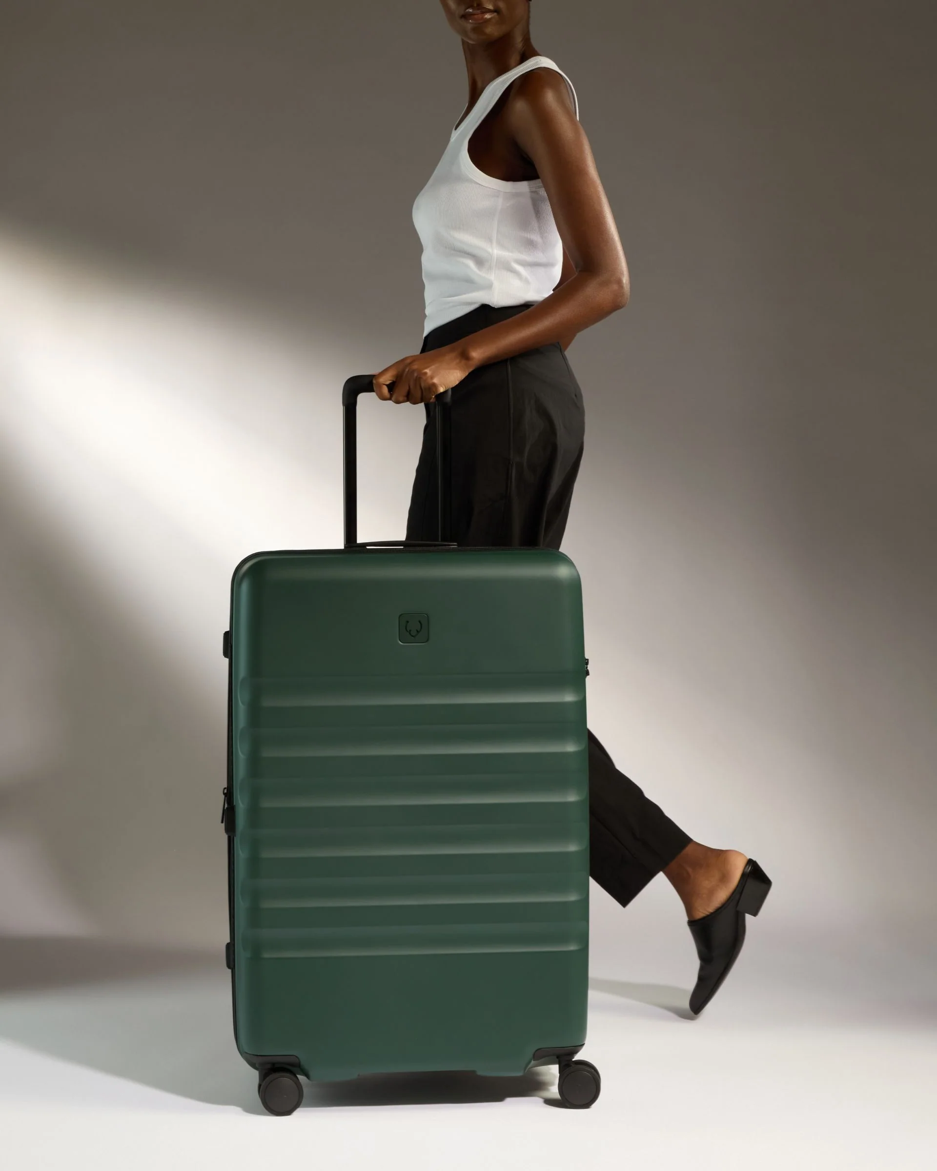Model walking with a green Antler suitcase in a studio travel scene.