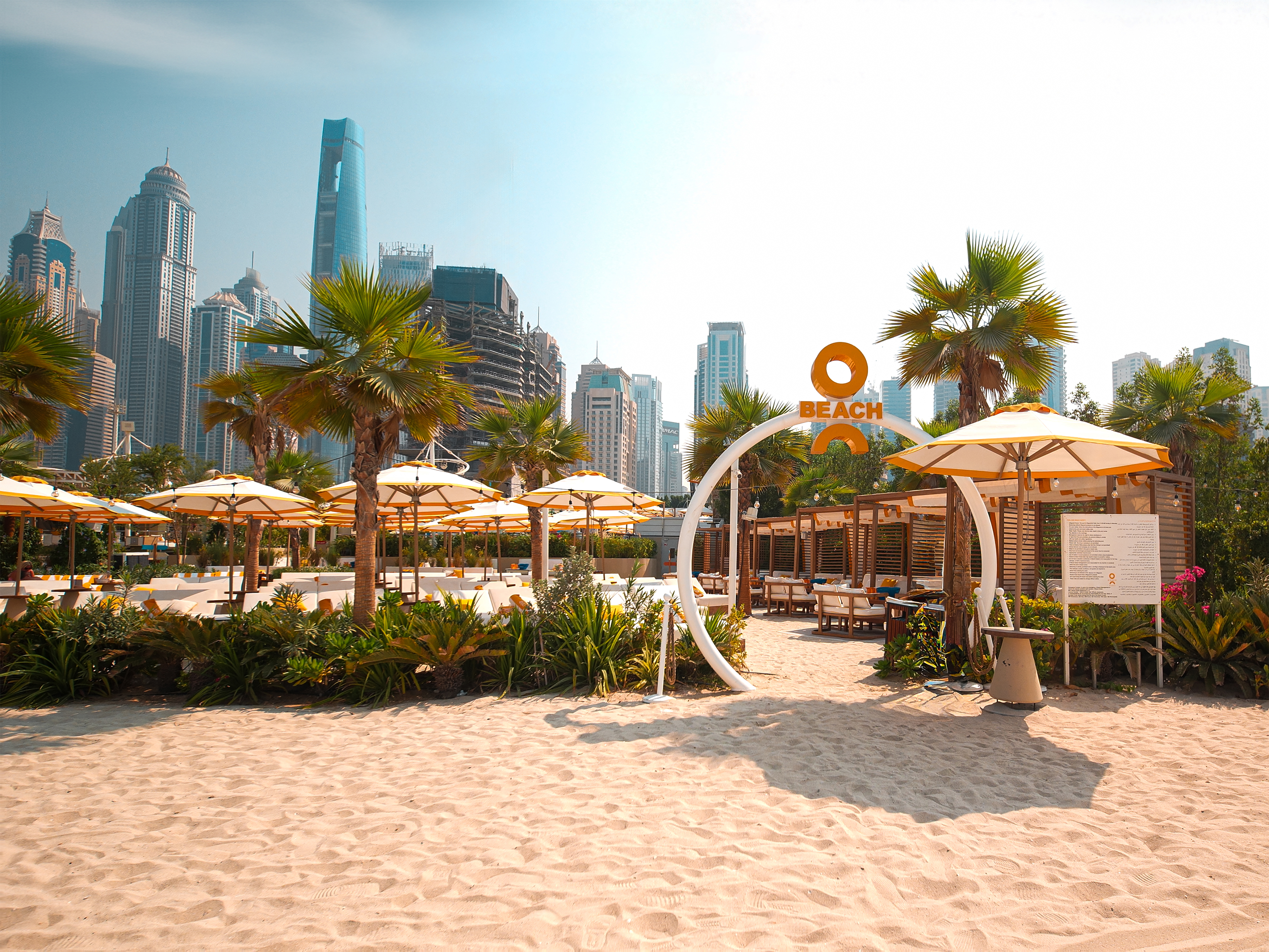 O Beach Dubai entry area with sand, palms, skyline, and brand sign.